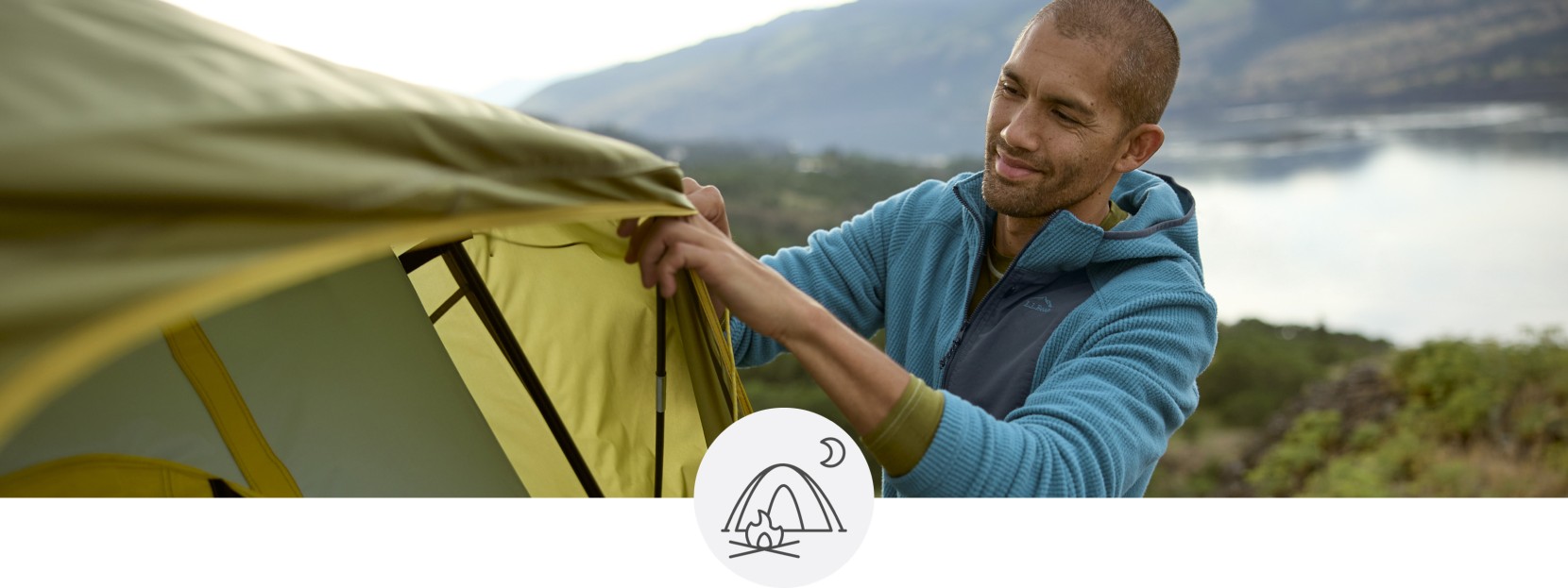 Close-up of a man setting up a tent, a lake and mountains in the background.