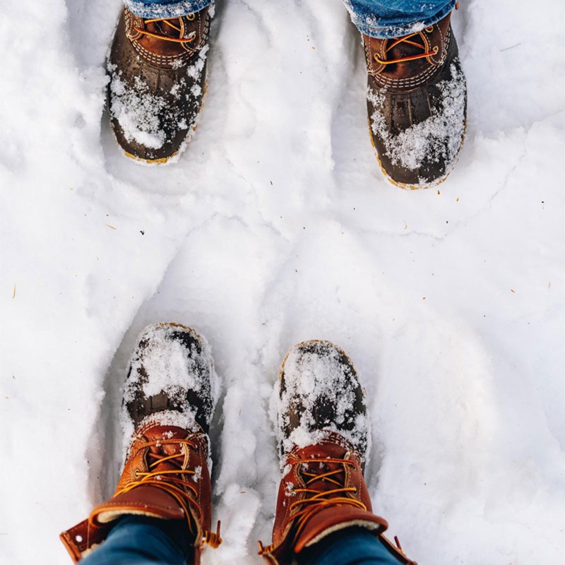 An overhead view of 2 sets of feet standing in snow wearing L.L.Bean Boots.