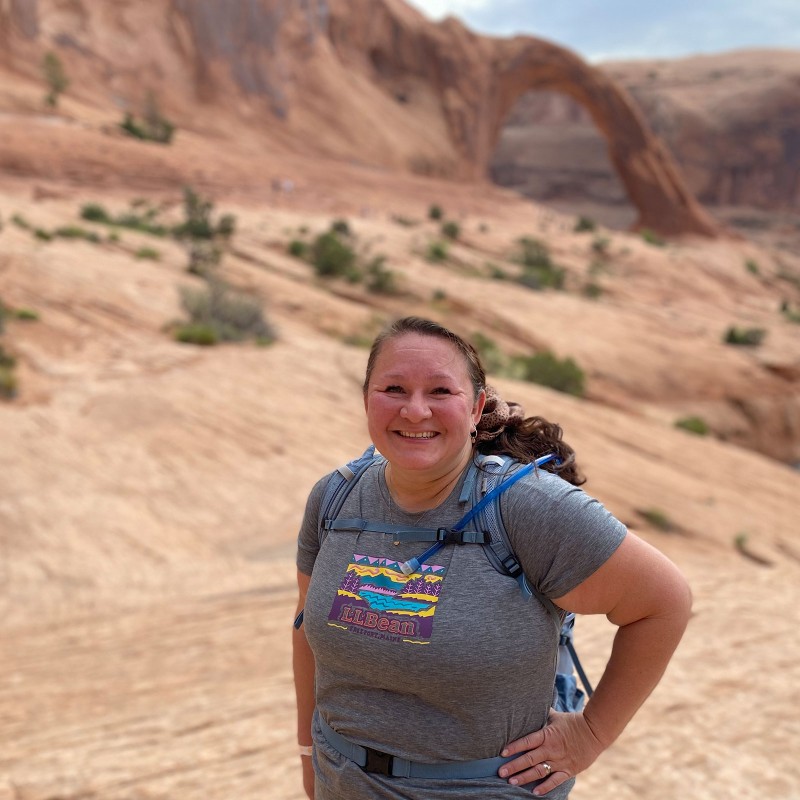 A smiling Melody Forsyth wearing a backpack standing in a desert-like landscape amidst rock formations.
