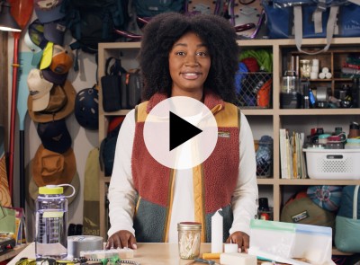 Stephanie standing at a table covered with camping essentials, camping and outdoor gear on shelves in the background, a play video icon in the center.