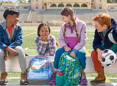 4 kids sitting on a bench on an athletic field with their school backpacks.