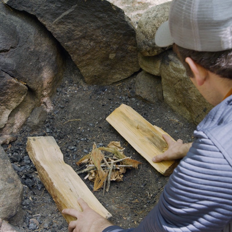 Nate placing split logs on either side of a pile of tinder in a fire ring.