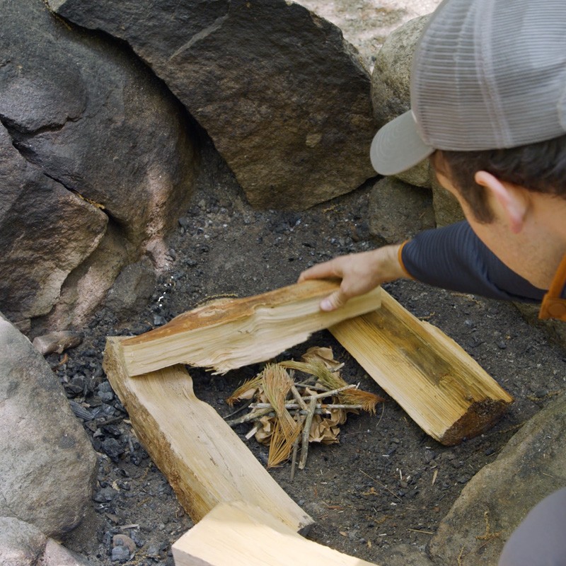 Nate placing 2 fuel logs crosswise on top of 2 existing logs with tinder in the center.