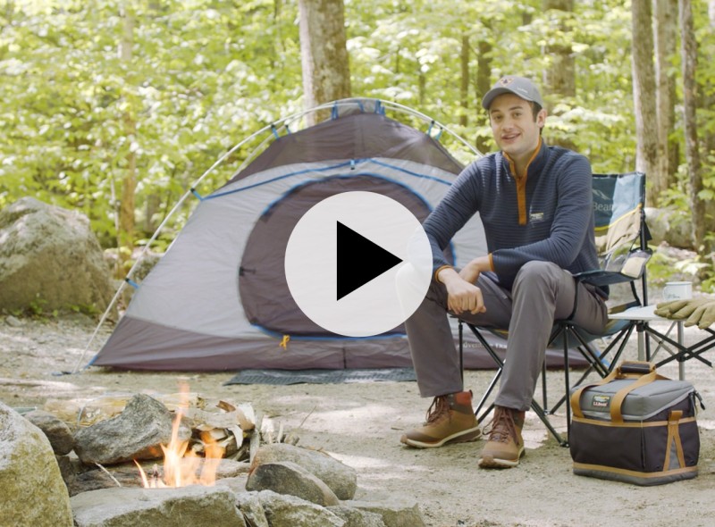 A man sits in front of a campfire in the woods next to a pitched tent in the woods and a play video icon in the middle.