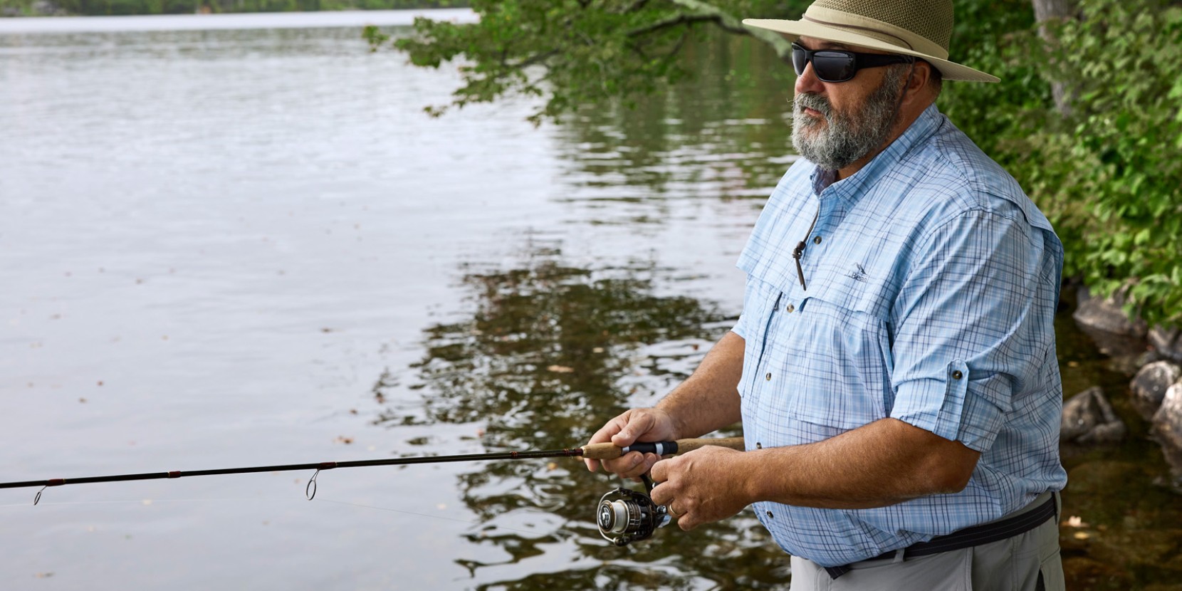 A man fishing from a river's edge wearing a wide-brimmed hat, sunglasses and a fishing shirt with sleeves rolled up.