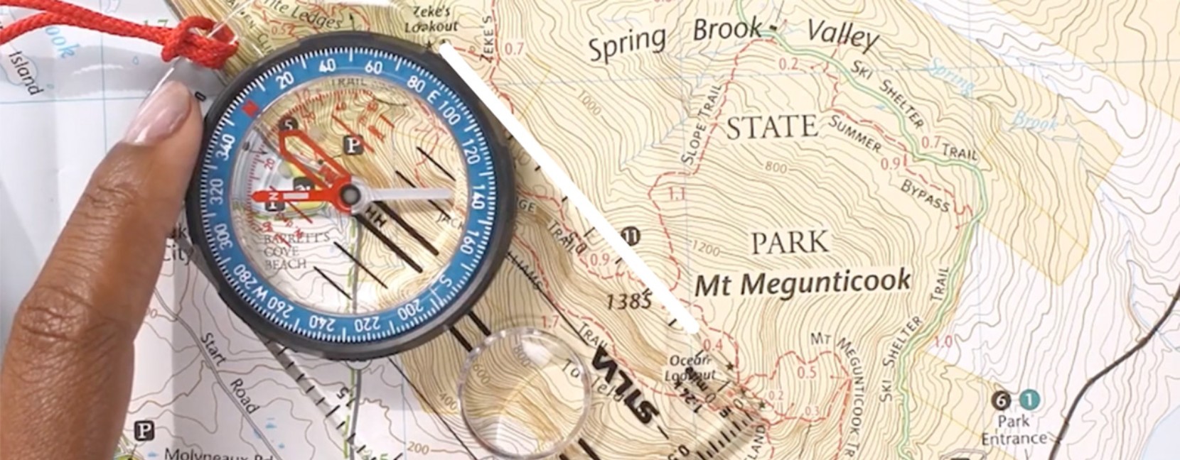 Close-up of a map with a compass on it, a white line indicates where the compass is aligned with the planned line of travel.