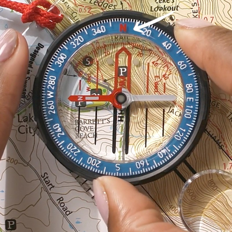 A close-up of fingers on the bezel of a compass on a map. A white arrow indicates