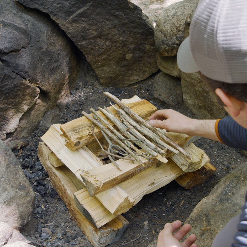 Nate laying twigs across the top of the log-cabin style fire as a "roof."