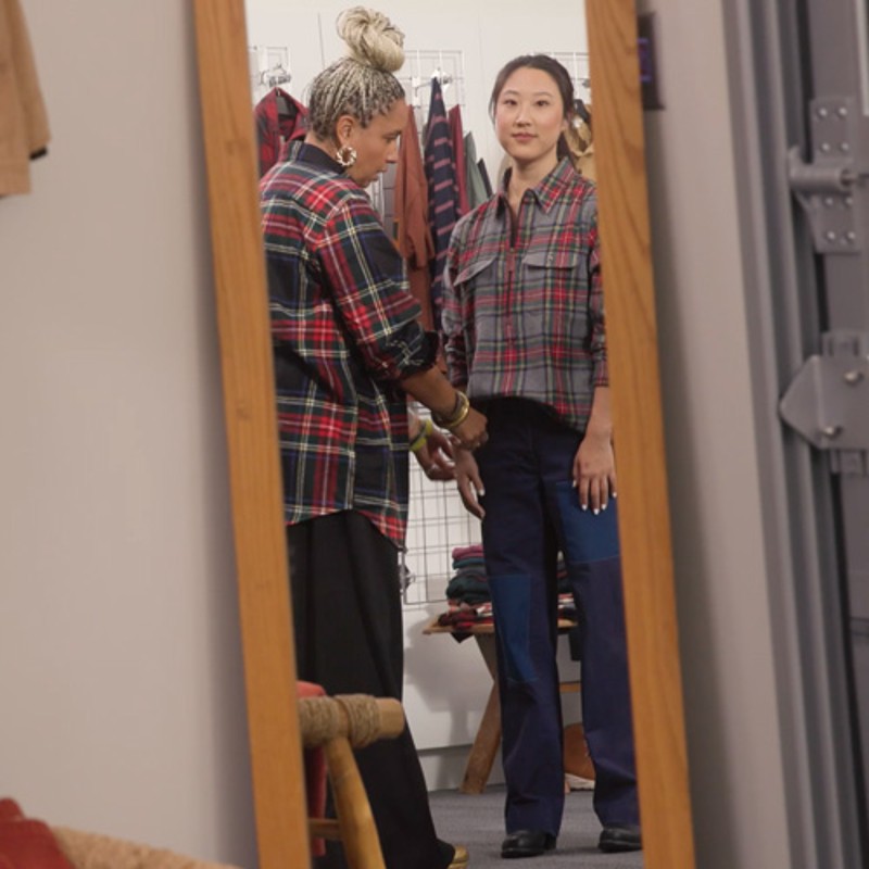 The stylist working with the sleeve of a flannel shirt worn by the model, reflected in a full-length mirror.