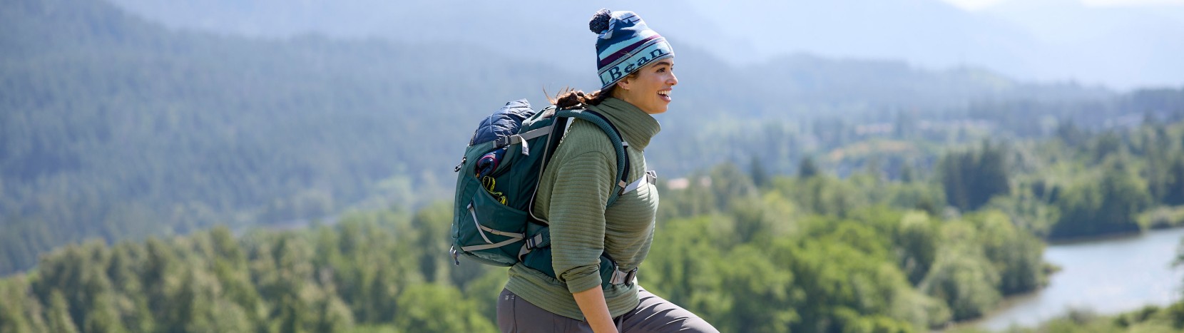Woman walking on a mountain trail wearing a backpack with a lake in the background