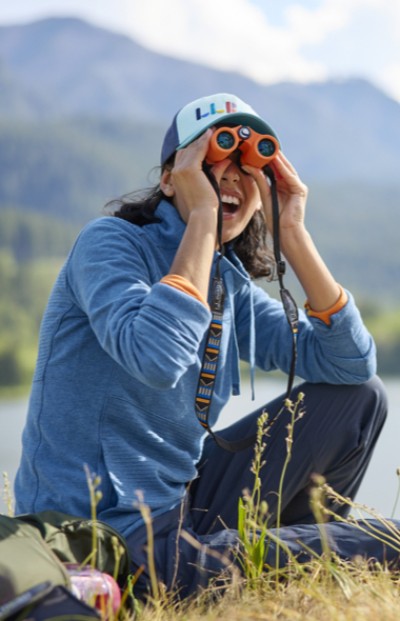 Image of a person sitting in the grass with a lake and mountains in the background using binoculars