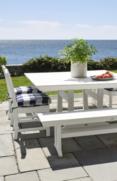 Image of white outdoor table and chairs with checkered pillows and side benches with a stone wall and ocean in the background