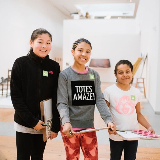 Three smiling kids stand inside a museum holding clipboards and papers.