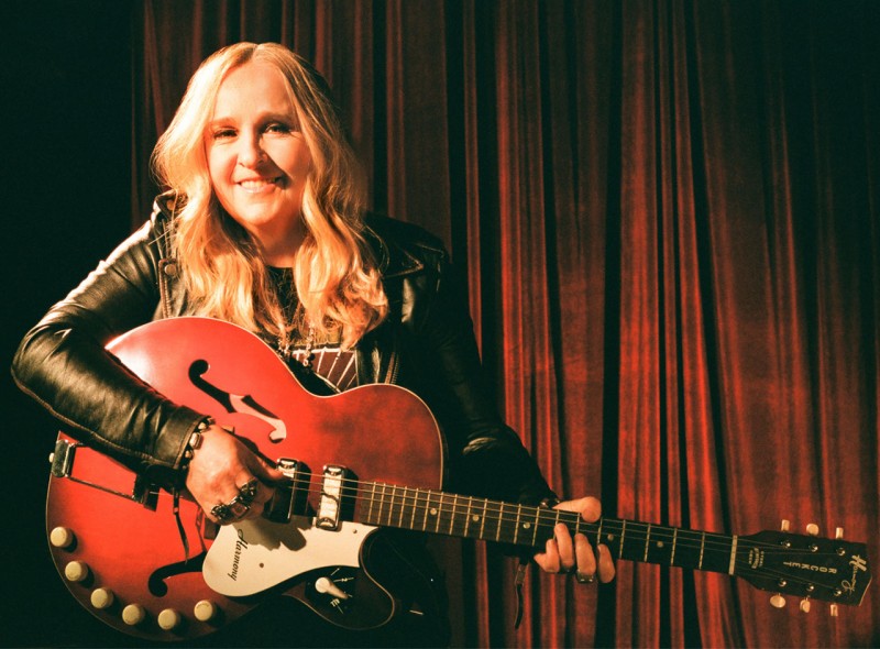 Melissa Etheridge with her guitar on stage, a drawn curtain behind her.