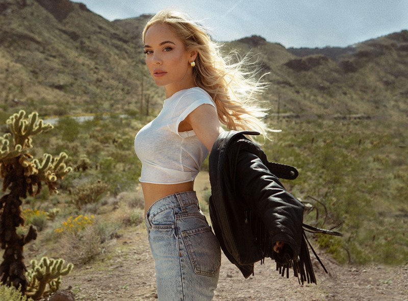 Mackenzie Porter standing sideways on a dusty desert trail, black leather jacket hanging on outstretched arms.