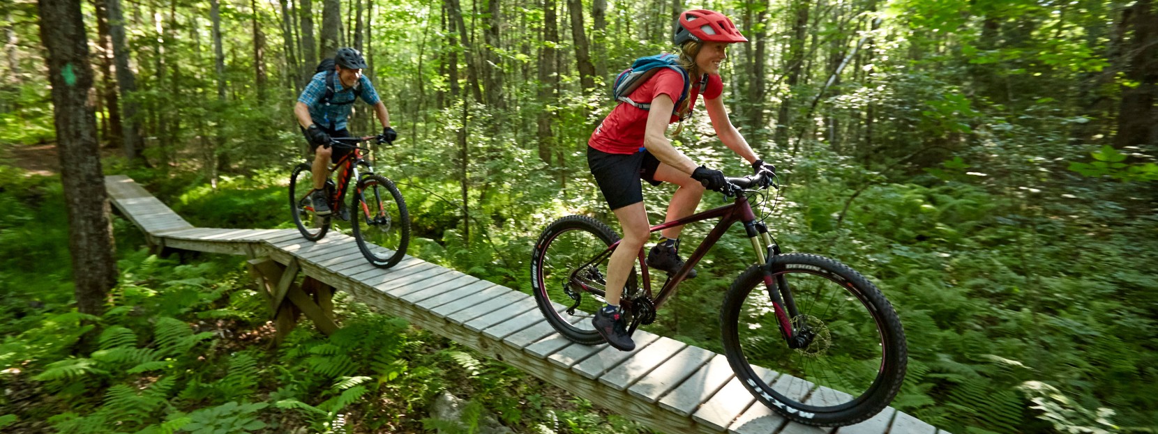 2 mountain bikers riding across a raised boardwalk in the woods.