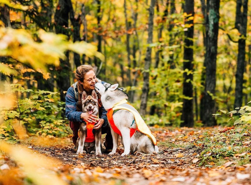 A woman scooching down to pet 2 dogs in the autumn woods.