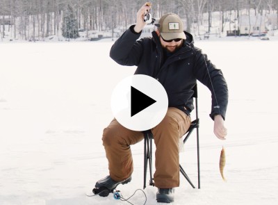 A man sits on a camp chair on the ice, pulling up a fish, a play video icon in the center.