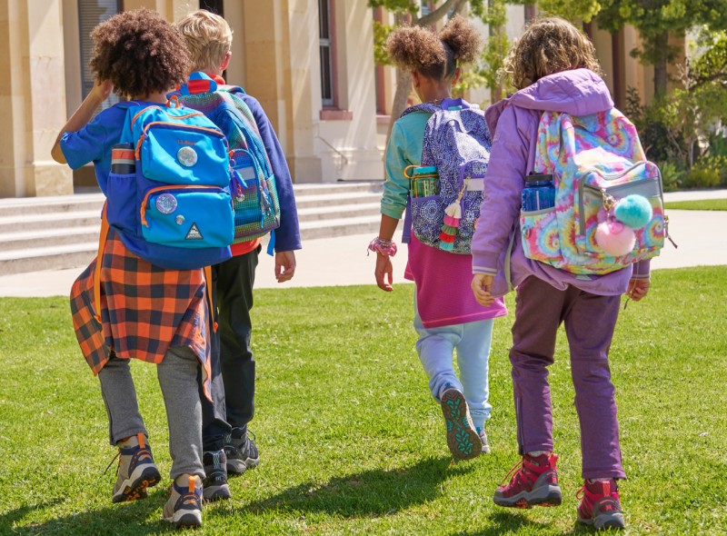 Several kids from behind walking into school with backpacks on.