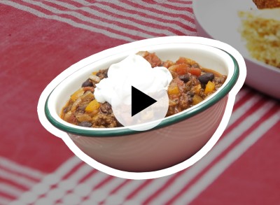A bowl of chili with a dollop of sour cream on a red and white checkered tablecloth with a play video icon in the center.