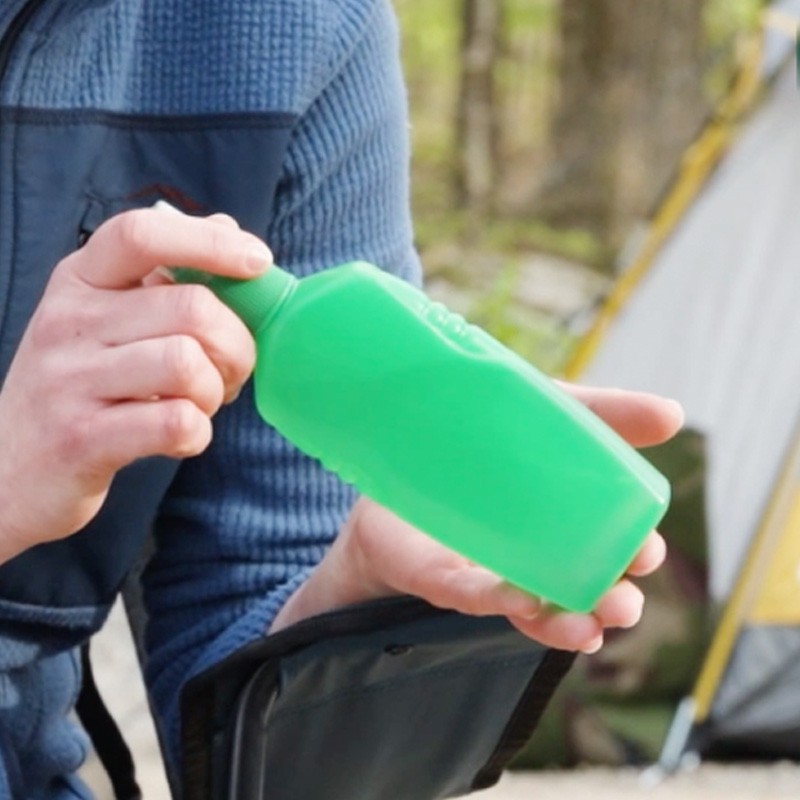 Hands hold a green bottle of bug spray with a tent in the background.