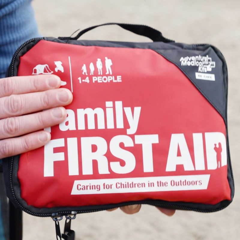 A hand holds a red Family First Aid kit, signaling preparedness for outdoor care emergencies. 