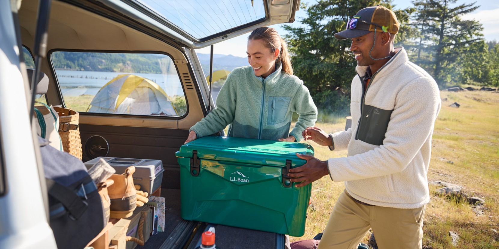 Two people unload a L L Bean cooler from a car near a tent by a lake.