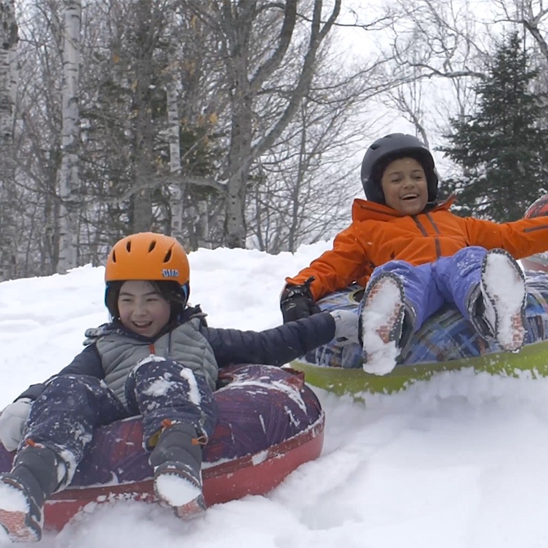 Children in a winter setting slide on snow using L L Bean Sonic Snow Tubes