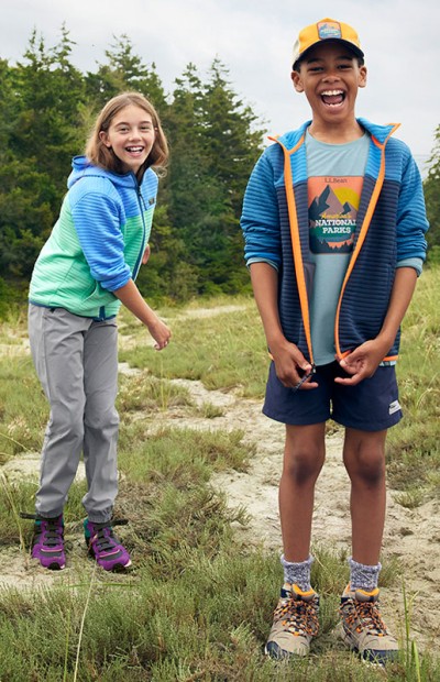 Two children wearing LL Bean outdoor clothing, smiling outside on a hiking trail.