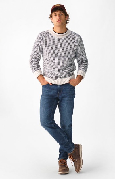 A man stands with one leg crossed over the other, hands in pockets, wearing a sweater, jeans, and brown shoes.