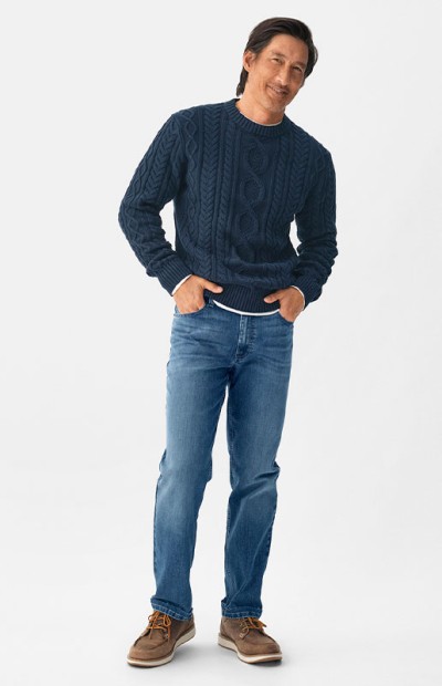 A person wearing a cable knit sweater, jeans, and boots stands with one hand in a pocket.