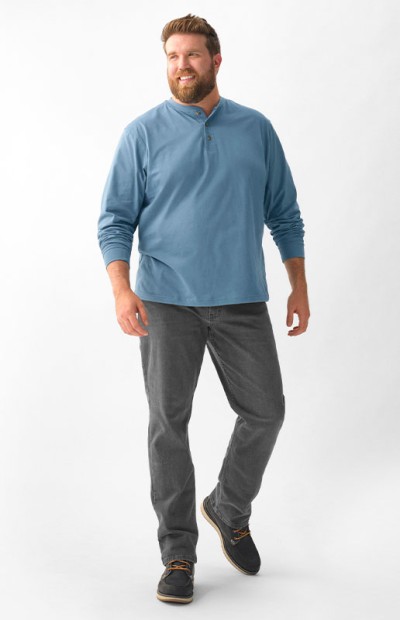 A man stands against a white background, wearing a blue top, grey pants, and brown shoes.