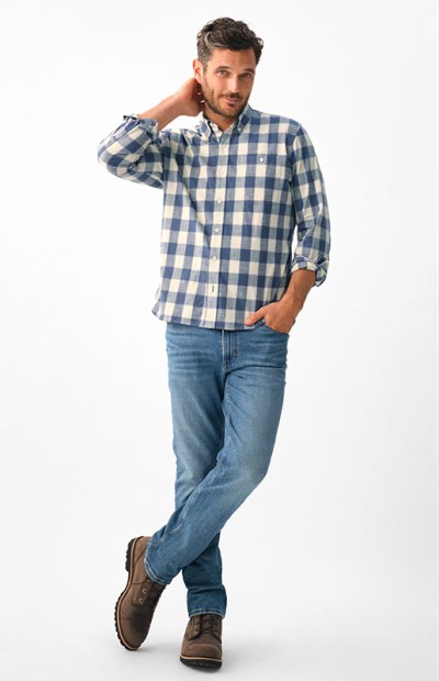 A man in a checkered shirt and jeans stands with one hand on hip, against a white background.
