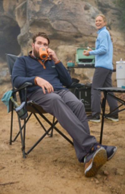 Man sitting in a camp chair, drinking coffee and woman enjoying camping with their camp kitchen.