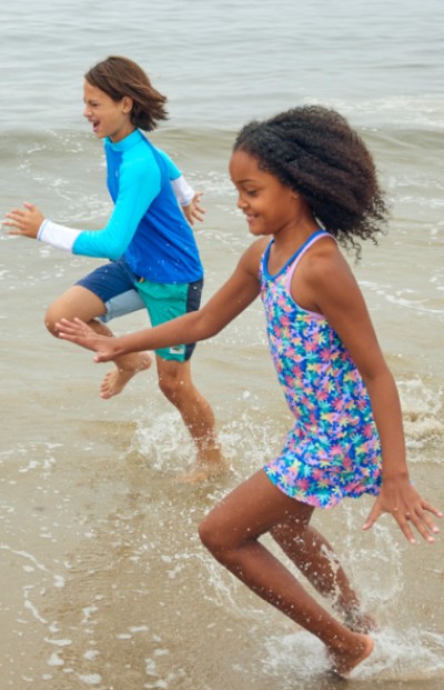 Two children wearing swimsuits running through the water at the beach.