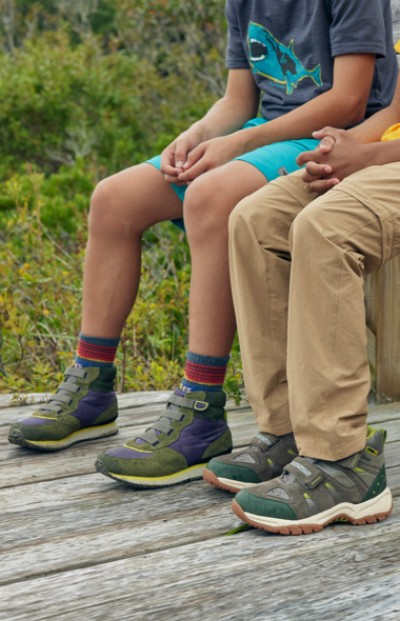 Two young kids wearing hiking boots sitting on a bench with wooded shrubs in the background.