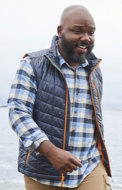 Man wearing a vest and flannel shirt walking by the ocean.