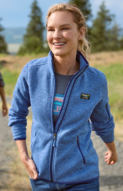 Woman wearing fleece top walking in a grassy field with pinetrees in the background.