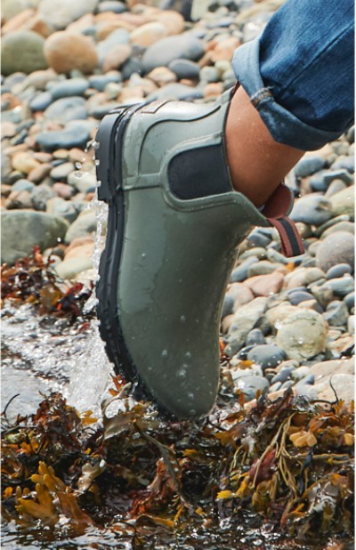 Image depicting a woman wearing rain boots walking on the rocky beach.