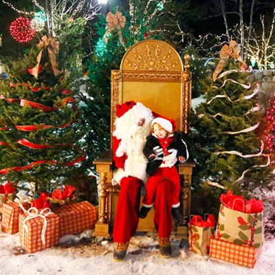 Santa sitting with a little one on his lap, surrounded by wrapped gifts and holiday trees.