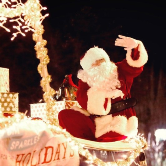 Santa, surrounded by wrapped presents, waving to a crowd from a brightly lit sleigh.