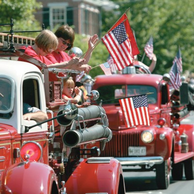 2 vintage fire trucks in the 4th of July parade carrying people waving American flags.