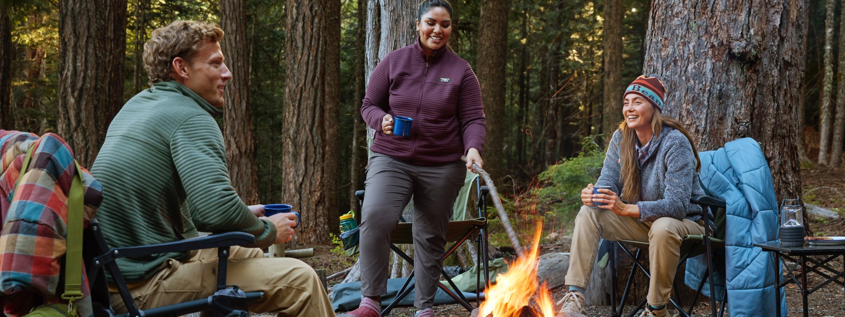 3 friends sitting around a campfire at their campsite, a wooded area in the background.