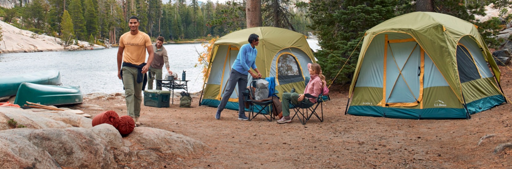 People setting up a lakeside campsite with tents, chairs, gear, and canoes among trees.