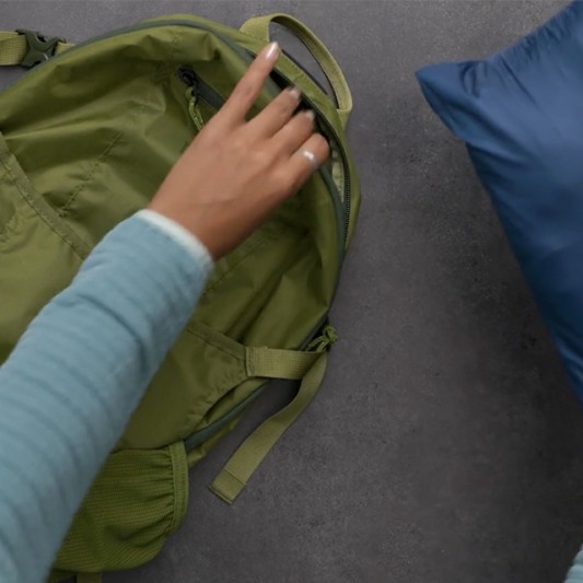 A hand opens a green backpack with a blue stowawayed jacket resting nearby