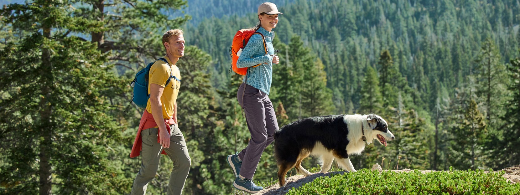 Two people with L L Bean backpacks and a dog hike on a trail in a forested area.