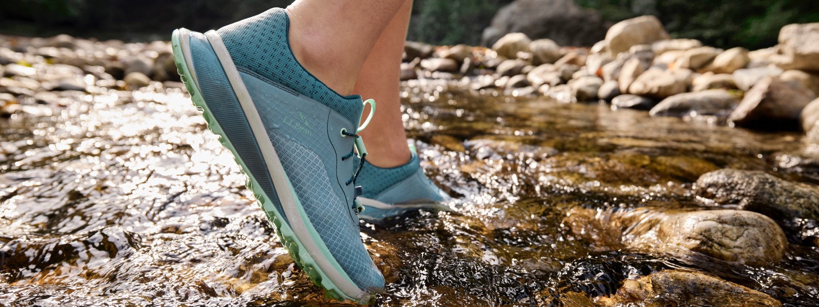 A person wearing blue athletic shoes with green soles stands in a shallow stream with visible rocks.