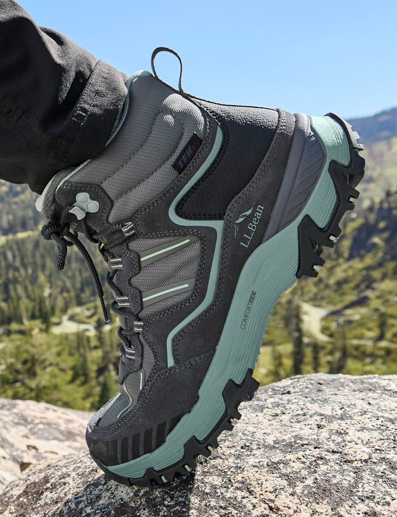 A grey and teal L L bean hiking boot is worn by a person standing on a rock.