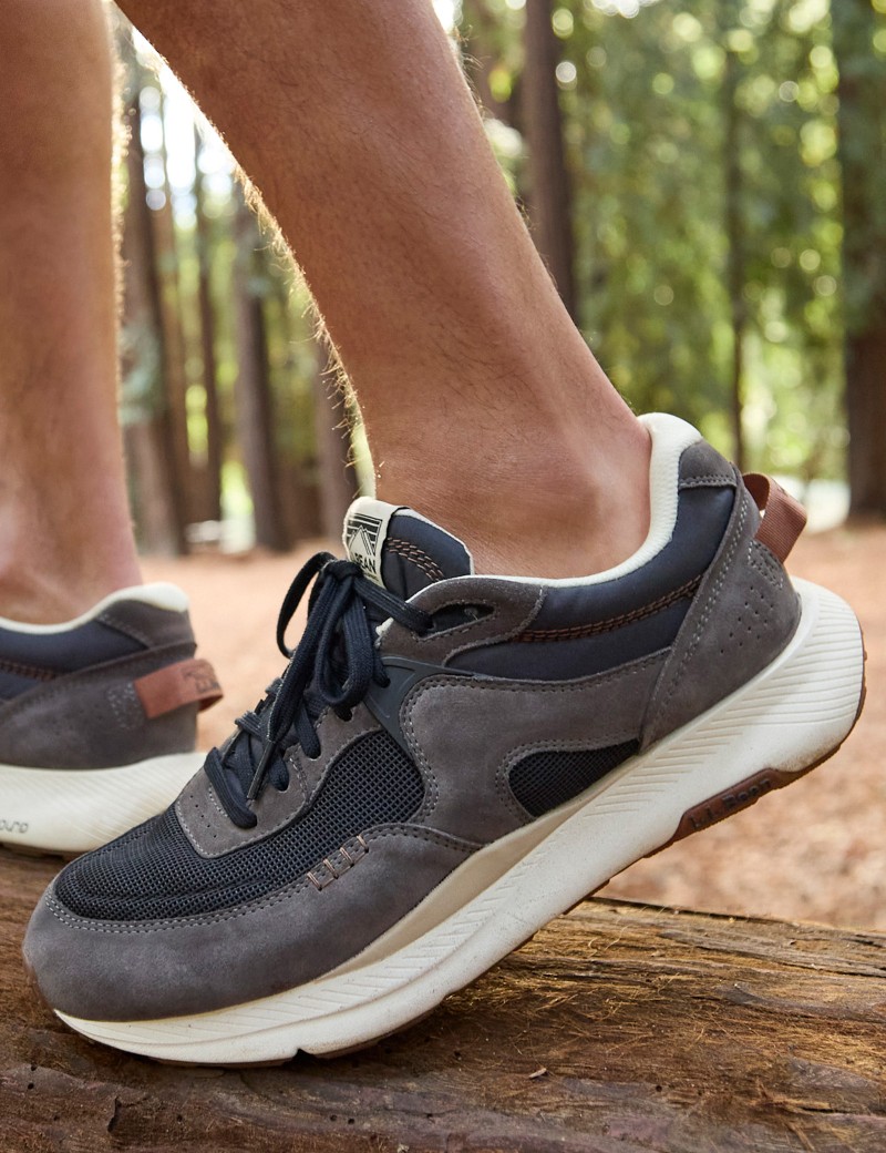 A person wearing gray and white sneakers with black laces stands on a log in a forest with tall trees. 
