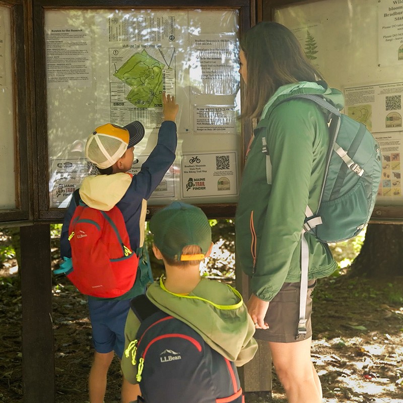 A mom with her two boys with backpacks looking at a trail map on a board in a forest.